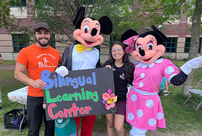 Two of our staff posing with life size disney characters showing off a sign that is decorated with flowers that says Bilingual Learning Center.
