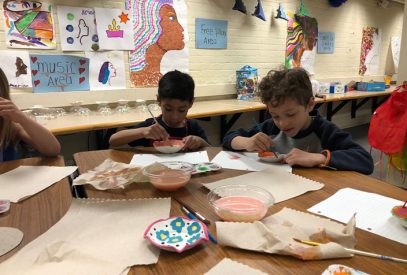 Two kids are focused on painting their clay bowls, with paintbrushes in hand and paints across the table. Different paintings are in the background.